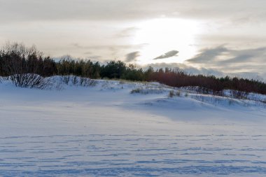 Güneş, karlı bir kıyı manzarasının üzerinde kum tepeleri ve çalılarla birlikte bir bulut katmanının arkasında batar. Sıcak altın ışık, beyaz kara huzurlu bir doğa görüntüsüyle vurur..