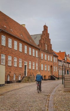 Viborg / Danimarka 'da bulutlu bir gökyüzünün altında tarihi tuğla binalarla dolu kaldırım taşlı bir sokakta bisiklet süren bir kadın..