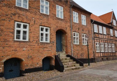 Danimarka 'da Viborg: Birden fazla beyaz çerçeveli pencereli tarihi tuğla binalar, mavi kapılar ve bulutlu bir gökyüzünün altındaki kaldırım taşı bir caddede taş basamaklar.