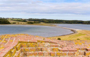 Danimarka 'da Kalo Slotsruin: Ön planda tuğla bir duvar ve arka planda sakin su ve yeşillik içeren eğimli bir kıyı şeridi bulunan parçalı bulutlu bir gökyüzünün altında manzaralı bir kıyı manzarası.
