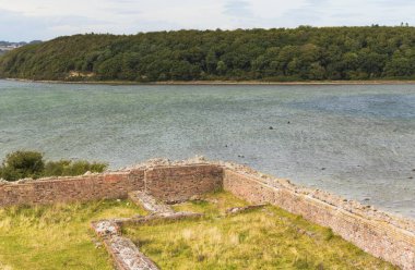 Danimarka 'daki Kalo Slotsruin: Bulutlu bir gökyüzünün altında arka planda ormanlık bir tepe olan bir su kütlesinin yakınındaki tuğla duvar kalıntıları.