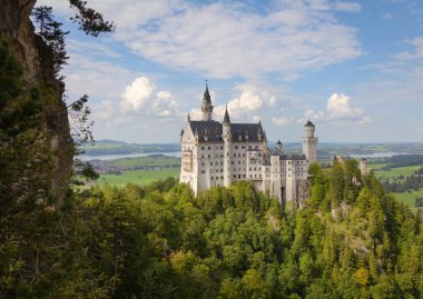 Neuschwanstein Şatosu parçalı bulutlu bir gökyüzünün altında manzaralı Bavyera kırsalına tünemişti..