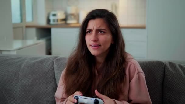 En flicka hemma sitter på soffan och spelar en konsol på en vit sladdlös joystick. Flicka förlorar och blir upprörd — Stockvideo