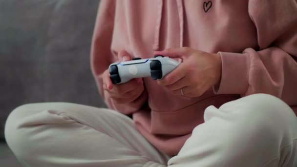 En tjej i rosa jacka och vita byxor hemma sitter på soffan och leker på en konsol på en trådlös joystick. Tryck aktivt på knapparna för att inte förlora — Stockvideo