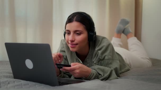 Das Mädchen liegt mit Kopfhörern hinter einem Laptop auf dem Bett, die Bankkarte in der Hand und füllt die Kartendaten aus. — Stockvideo
