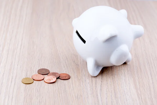 White piggy bank on a wooden desk: it's on its side with a few penny's, a concept of a tight budget