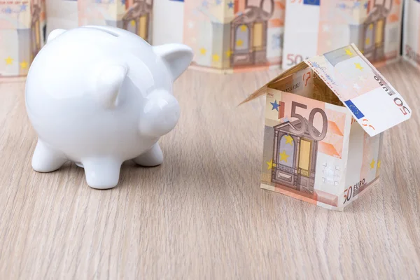 Bank note house with white piggy bank on desk - real estate, home savings, mortgage, rent concept