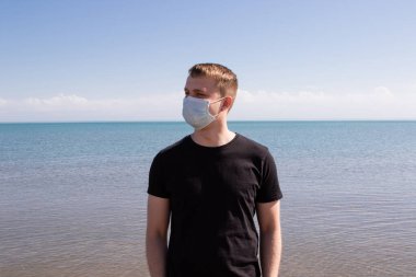 Mavi tıbbi maskeli bir adam denizde duruyor. Karantina sırasında dinlenin.