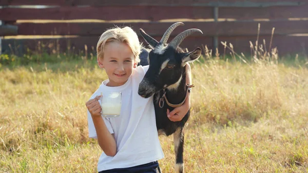 A boy drinks goat milk from a mug next to his goat.