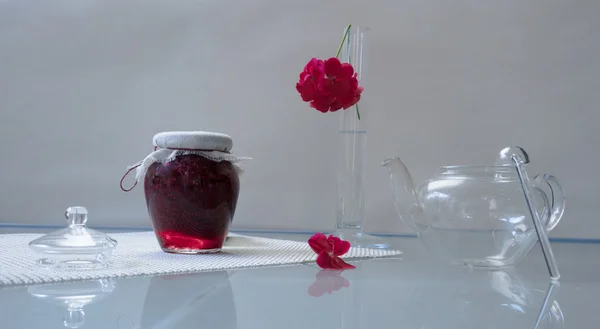 Образ жизни. Банк малинового варенья с чаем и цветком пеларгония на стеклянном столе . — стоковое фото