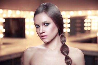 Beauty portrait of girl with beautiful makeup and long braid hair in dressing room. Showroom lamps. clipart