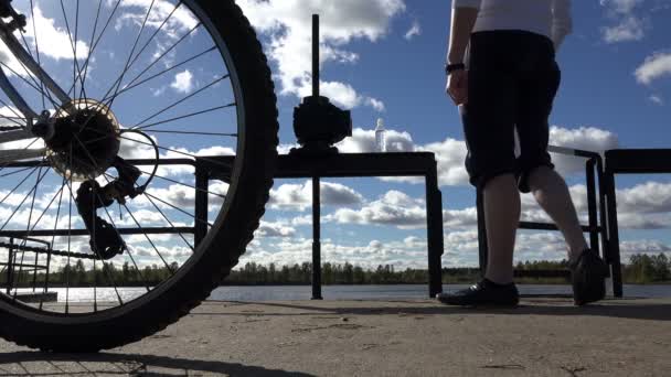 Усталый велосипедист пьет воду из бутылки, чтобы остаться в лагере рядом с плотиной на фоне голубого неба и головы велосипед недели — стоковое видео