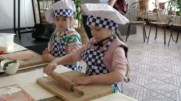 Dos niñas vestidas con uniformes chef aprendiendo a desplegar la masa con un rodillo en una tabla — Vídeo de stock