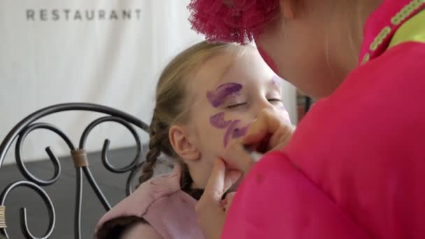 Professionele make-up artiest voert akvagrim opvallend paars kwast op het gezicht van een kind draagt de vlinder patroon — Stockvideo