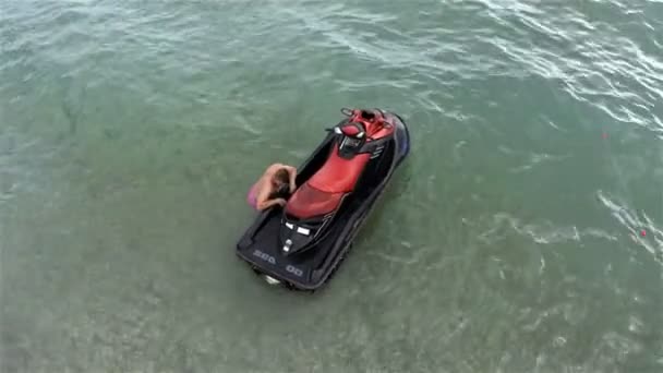 En ung man står midjan djupt i havet bredvid en jet ski — Stockvideo