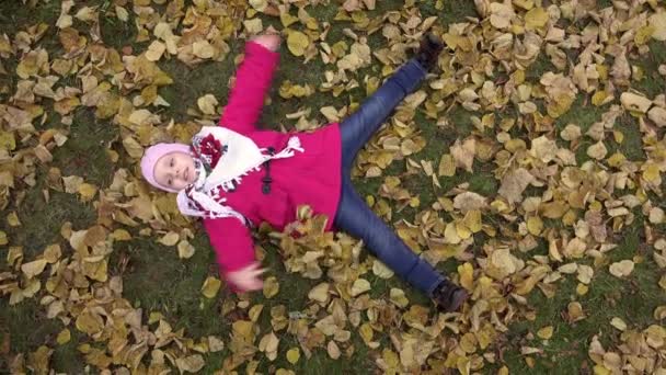 Menina deitada na grama com folhas acenando seus braços e pernas — Vídeo de Stock
