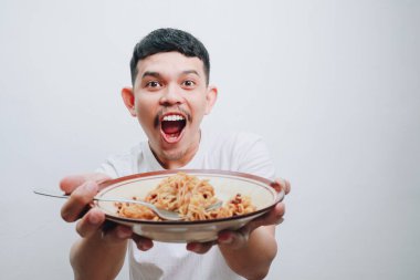 Neşeli genç bir adam büyük bir gülümsemeyle bir tabak erişte gösteriyor, heyecanını ifade ediyor..