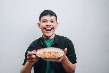 Genç bir adam neşeyle elinde bir kase erişte tutuyor, parlak gülümsemesini sergiliyor..