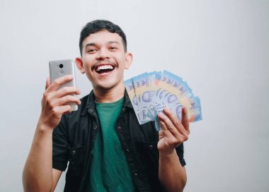Mutlu bir genç adam neşeli bir ortamda elinde para ve akıllı telefon tutarken gülümser..