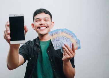 Neşeli genç bir adam heyecanlı bir şekilde akıllı telefonunu ve parasını gösteriyor..