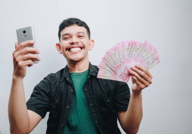 Neşeli genç bir adam elinde akıllı bir telefonla gururla para gösteriyor, neşe saçıyor..