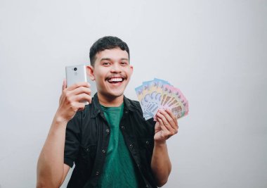 Neşeli bir genç adam gururla para ve akıllı telefon sergiliyor, başarıyı simgeliyor..