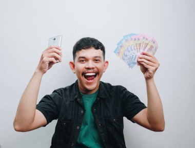 Neşeli genç bir adam elinde akıllı telefon ve nakit parayla başarıyı kutluyor..