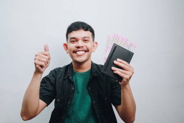 Neşeli bir genç adam gülümserken ve başparmağını kaldırırken nakit para ve defter sergiliyor..