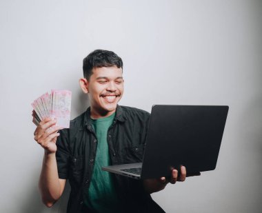 Neşeli bir genç adam nakit para tutuyor ve minimalist bir ortamda dizüstü bilgisayar kullanıyor..