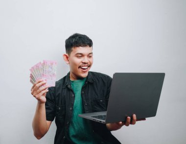 Genç bir adam dizüstü bilgisayarını kullanarak, başarıyı ve heyecanı somutlaştırırken neşe içinde para tutuyor..