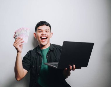 Neşeli gülüşü olan genç bir adamın elinde para ve dizüstü bilgisayar var..
