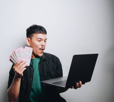 Genç bir adam elinde para tutarken ve dizüstü bilgisayar kullanırken şaşkınlığını dile getiriyor..