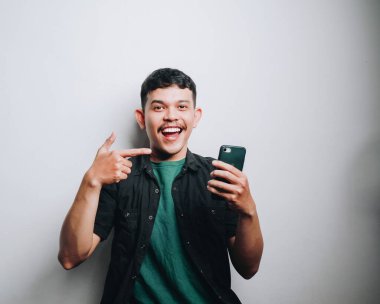 Neşeli genç bir adam, parlak bir ortamda gülümserken telefonunu işaret eder..