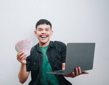 Neşeli bir genç elinde para ve dizüstü bilgisayarla mali başarısını kutluyor..