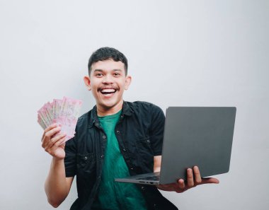 Neşeli genç bir adam para sergilerken ve dizüstü bilgisayarı üzerinde çalışırken finansal başarısını kutluyor..