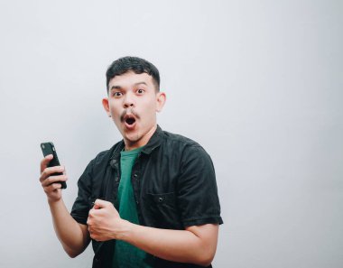 Şaşırmış bir adam telefonunu tutarken sevinçle tepki veriyor, heyecanını dile getiriyor..