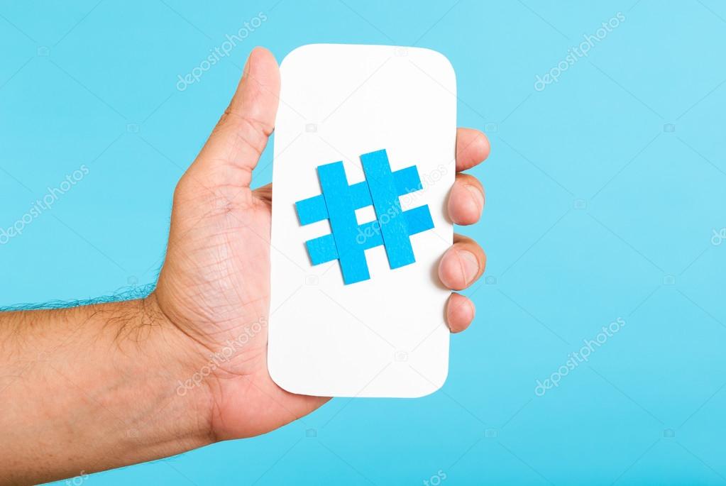Hand showing a hashtag symbol / sign on white paper with phone mobile shape, with blue background. Internet, social media concept.