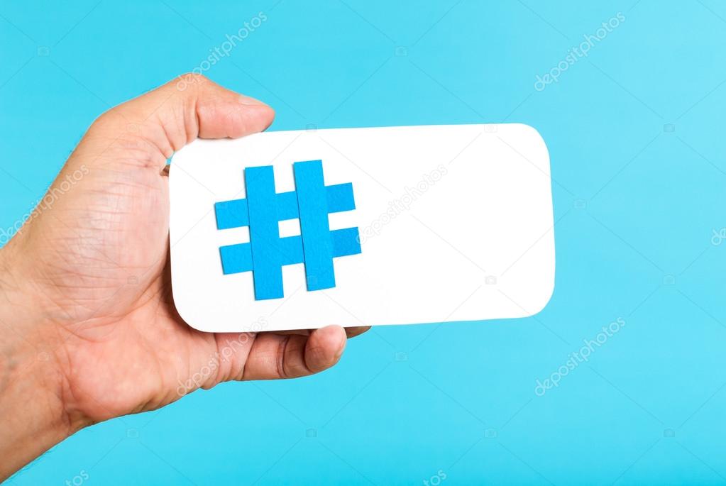 Hand showing a hashtag symbol / sign on white paper with phone mobile shape, with blue background. Internet, social media concept