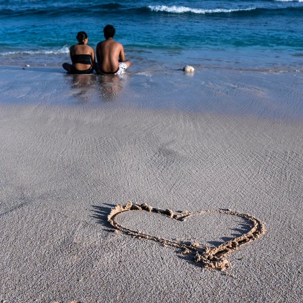 Lovely couple sitting on the beach and enjoing the sea and hLovely couple sitting on the beach and enjoying the sea and heart drawn on a sand in foregroundeart — Stock Photo, Image