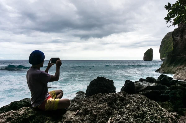 Jeune homme assis sur le rocher et faisant une photo des vagues sur smartphone — Photo