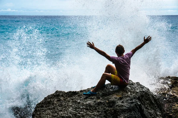 Jeune homme avec les mains levées assis sur le rocher avec les vagues de la mer en face Images De Stock Libres De Droits