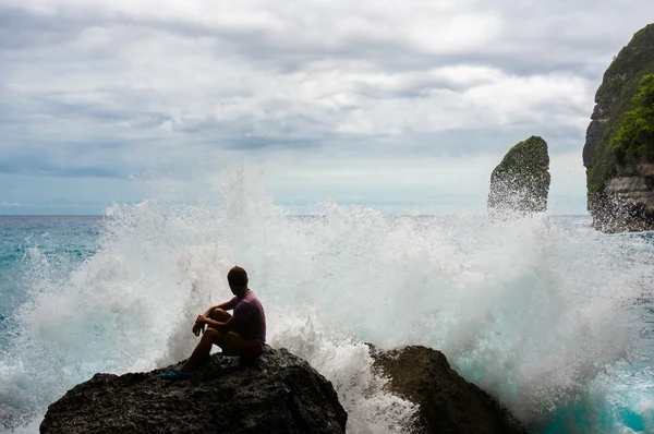 Молодой человек, сидящий на скале с морскими волнами, разбивающимися впереди — стоковое фото