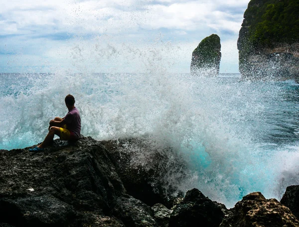 Jeune homme assis sur le rocher avec les vagues de la mer se brisant en face Photo De Stock