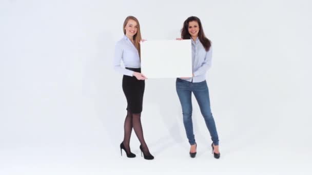 Dos chicas sonrientes caminando y sosteniendo una pancarta blanca con copyspace para publicidad o texto — Vídeos de Stock