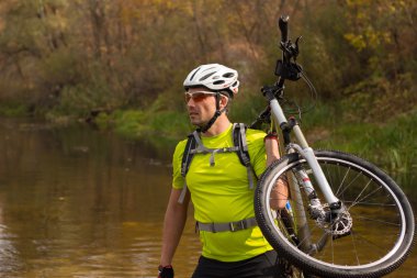 Bir adam bir kask ve bir sırt çantası onun omzuna bir bisiklet ile bir nehir geçer.