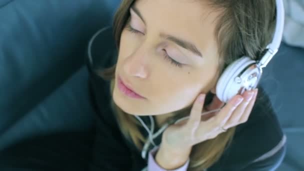 Hermosa chica disfruta escuchando música en los auriculares mientras está acostada en el sofá — Vídeo de stock