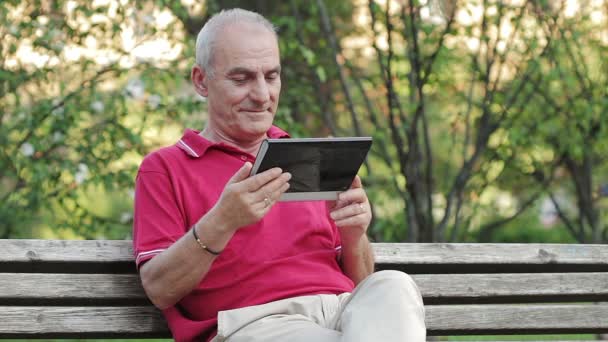 Пенсионер отдыхает и пользуется планшетом за столом в парке — стоковое видео