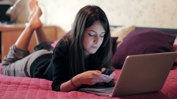 Chica usando el teléfono móvil en la cama mientras navega por Internet con el ordenador portátil — Vídeo de stock