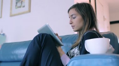 tablet pc ve Kupası evde kanepenin üstünde oturan genç kadın