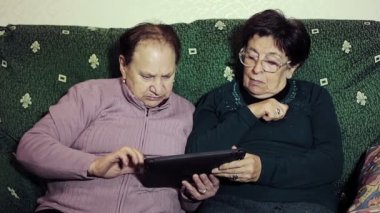 Yaşlı kadınlar evde birlikte oturup tablet bilgisayar kullanıyorlar.